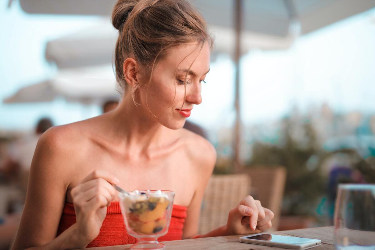 Chica sentada en la terraza de un restaurante comiendo un postre y pagando la cuenta con su teléfono móvil