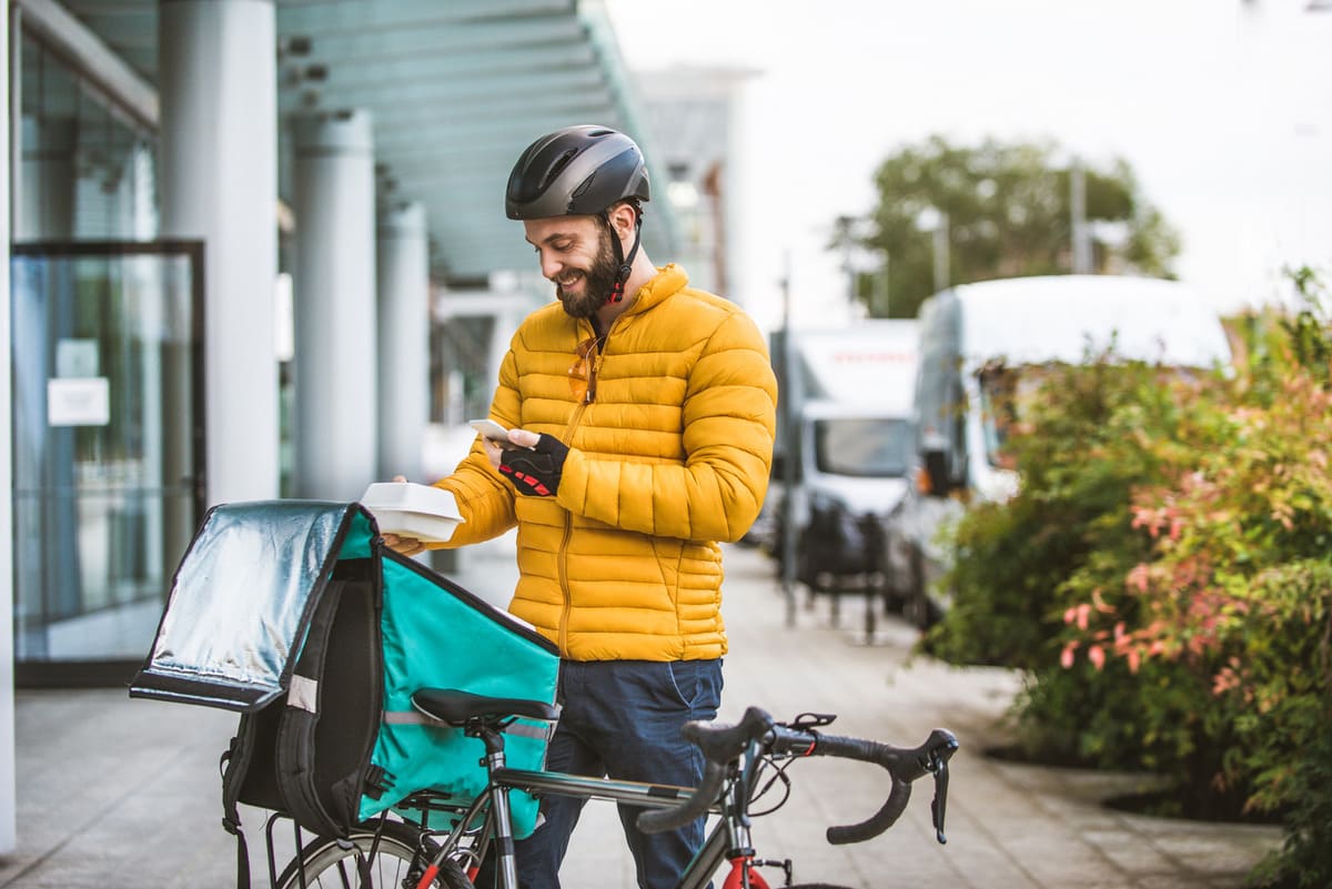Repartidor de comida a domicilio con su bicicleta sacando los productos de la mochila isotérmica y mirando el móvil