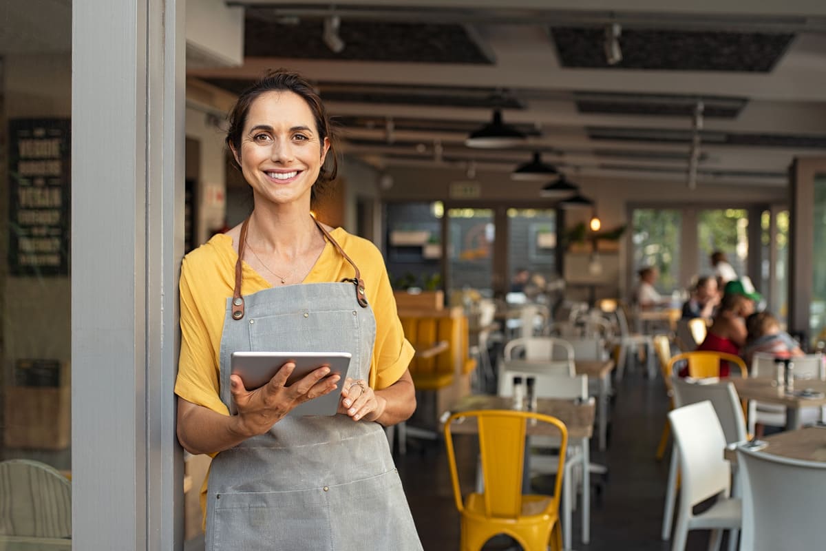 Camarera sonriendo apoyada en la puerta de un restaurante con un tablet en la mano